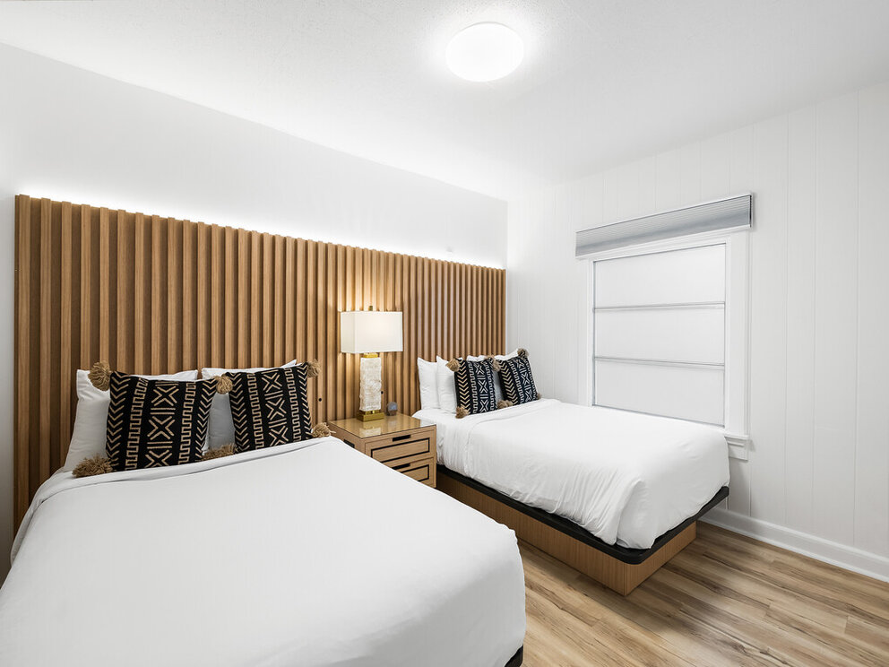 A clean and modern hotel room with two twin beds, a wooden headboard, and a nightstand with a lamp.