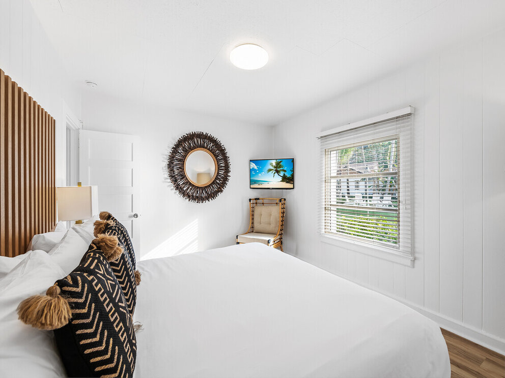 A white guest room with a large bed, wood headboard, and window with blinds.