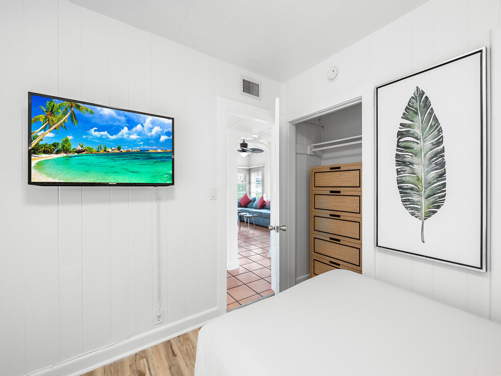 A white-walled guest room with a bed, TV, closet, and doorway leading to another room.