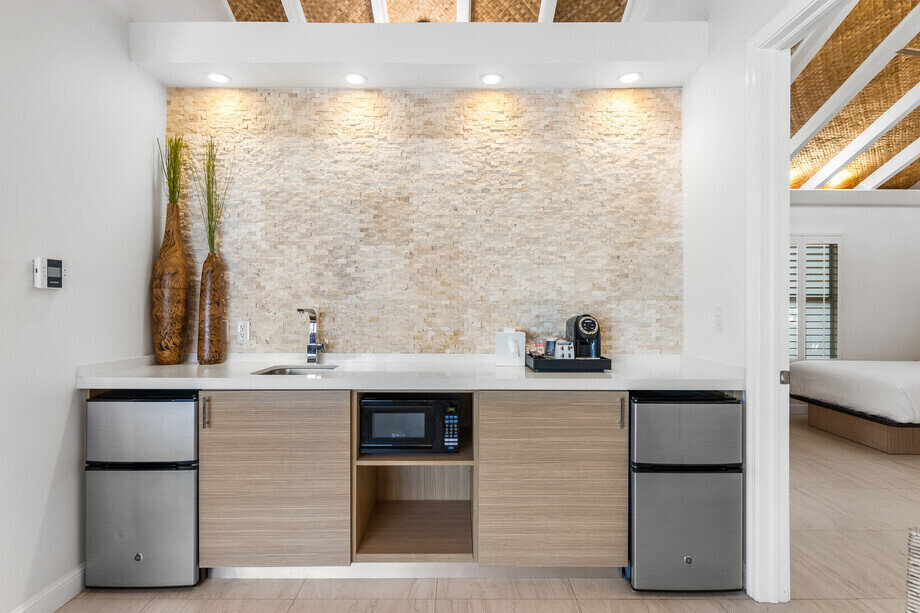 A modern kitchenette with a sink, microwave, mini-fridges, and coffee maker, adjacent to a bedroom.